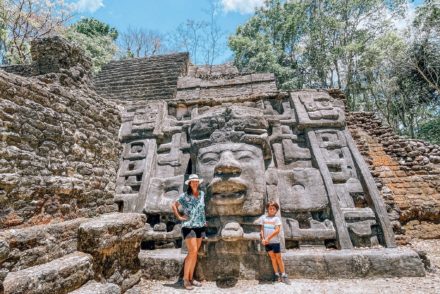 A woman and a boy standing in front of the Lamanai ruins in Belize. There is a gorgeous stone temple with a large carved face.