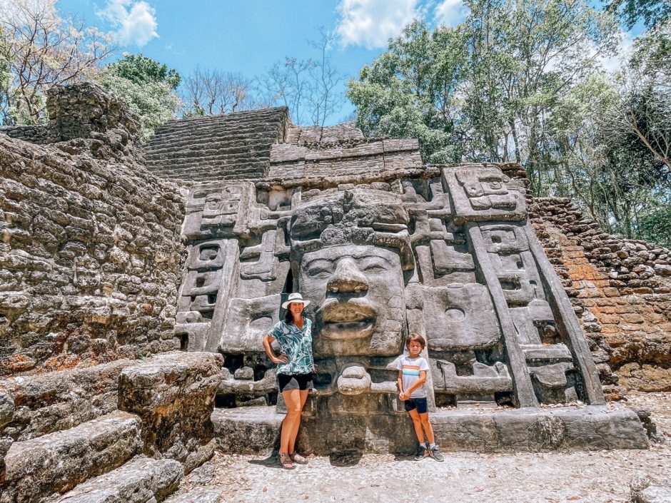 A woman and a boy standing in front of the Lamanai ruins in Belize. There is a gorgeous stone temple with a large carved face.
